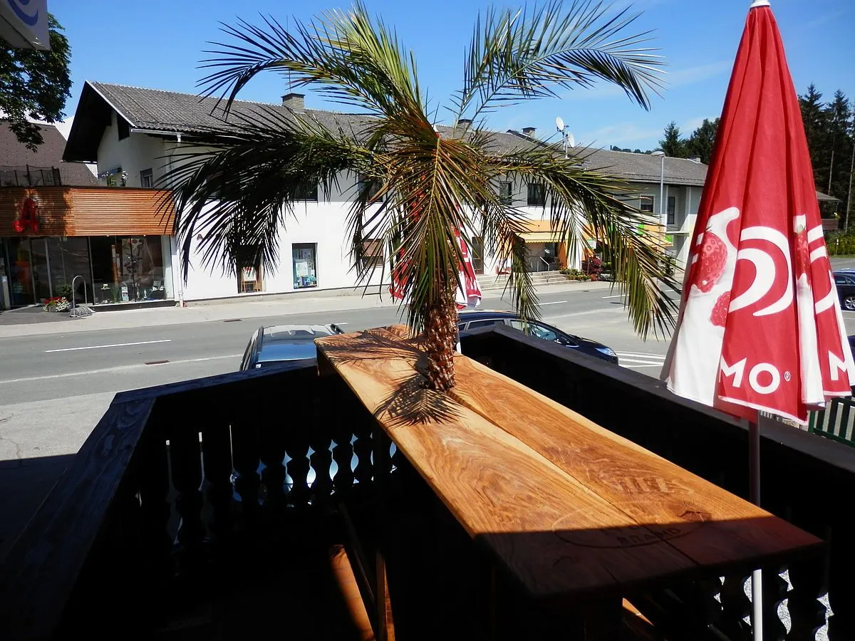 Ein Balkonblick mit einer Palme auf einem Holztisch. Zwei Autos sind auf der Straße geparkt, und ein Gebäude ist im Hintergrund. Der Himmel ist klar.