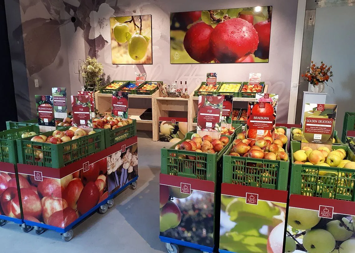 Ein Obst- und Gemüsebereich mit verschiedenen Kisten voller Äpfel, einschließlich Fuji und Braeburn, auf Rädern angeordnet, mit Wandpostern darüber.