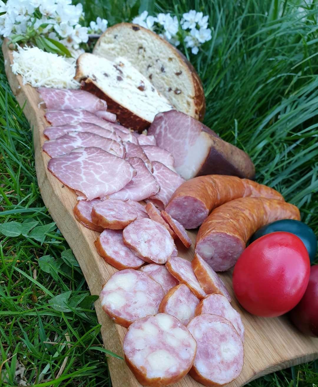 Ein Holzbrett zeigt eine Auswahl an Fleischsorten, darunter geschnittener Schinken, Würste und Salami, auf einer Rasenfläche neben bunten Eiern und einem Stück Brot mit Käse.
