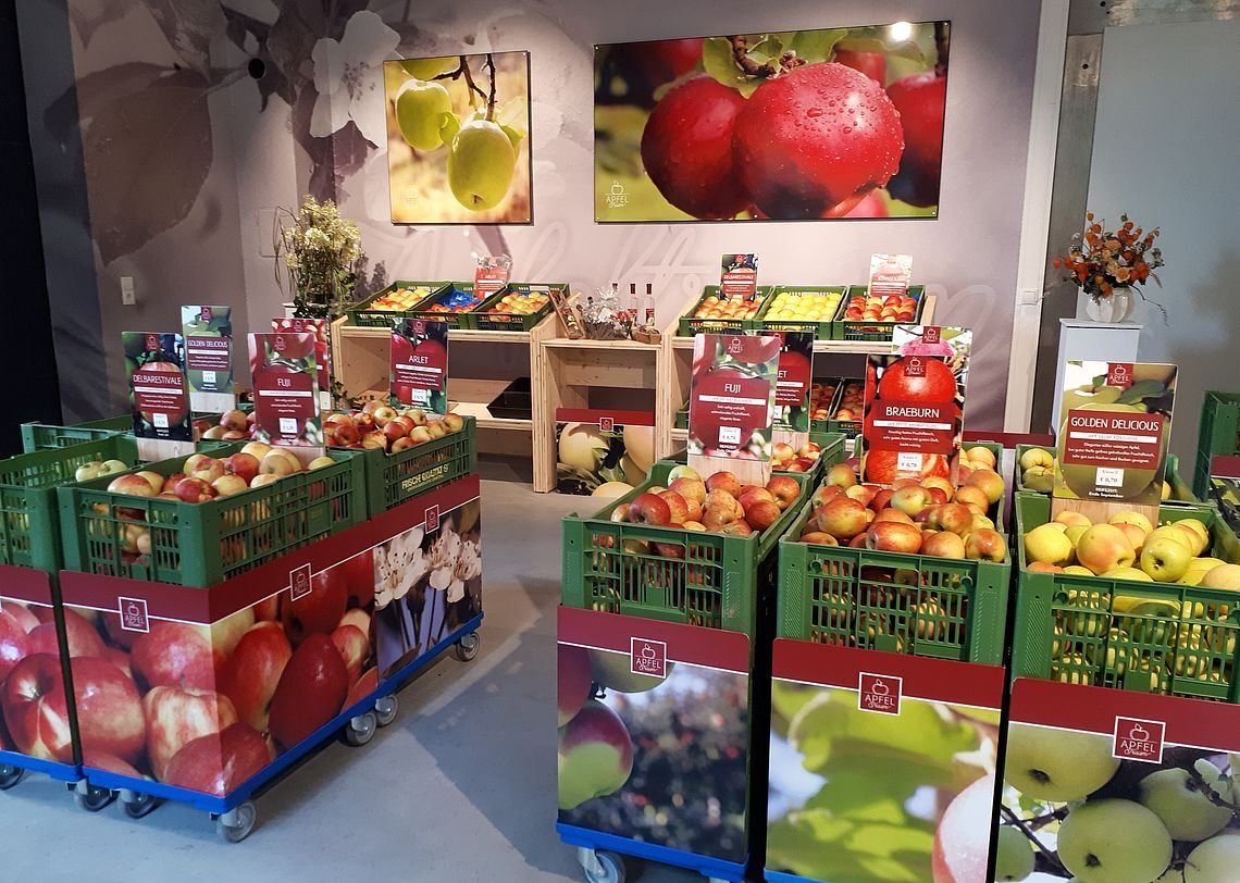 Ein Obst- und Gemüsebereich mit verschiedenen Kisten voller Äpfel, einschließlich Fuji und Braeburn, auf Rädern angeordnet, mit Wandpostern darüber.