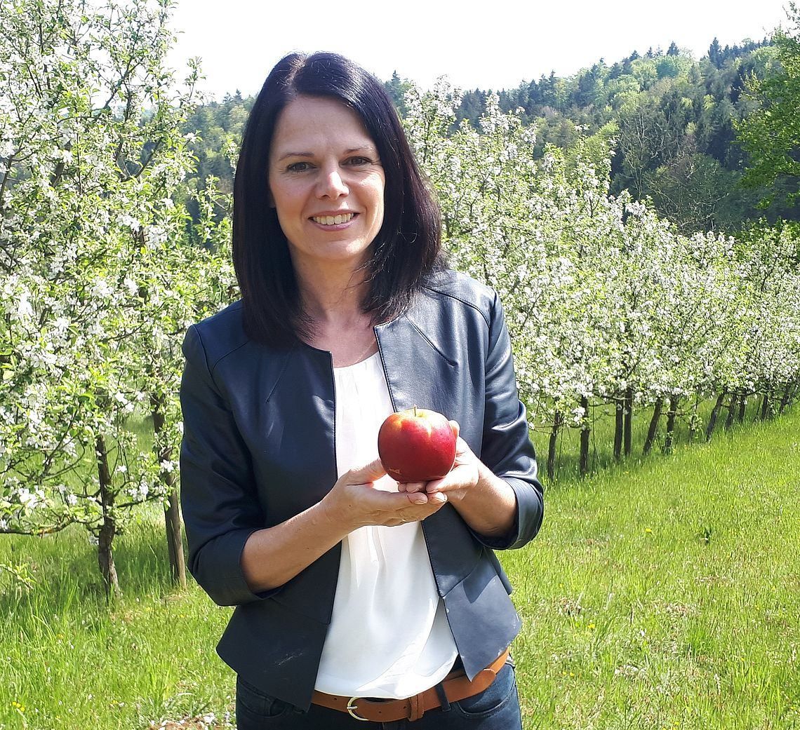 Eine Frau steht in einem Apfelhain und hält einen roten Apfel in den Händen. Sie lächelt und ist in einem weißen Hemd, einer marineblauen Jacke und Jeans gekleidet. Hinter ihr säumen Apfelbäume den Weg, und der Himmel ist klar.