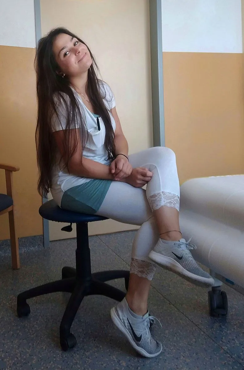 Eine junge Frau sitzt auf einem Stuhl in einem Raum. Sie trägt ein weißes Shirt, bunte Leggings und Nike-Sneaker. Sie lächelt und wirkt entspannt.