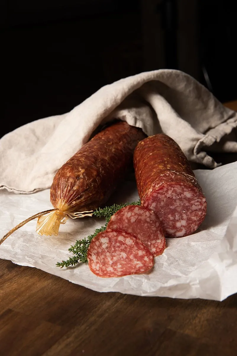 Zwei Scheiben Salami liegen auf einem Stück Papier, eine teilweise von einem Tuch bedeckt. Daneben befindet sich ein Bündel Salami mit einem gelben Faden. Ein Rosmarinzweig ist ebenfalls sichtbar.