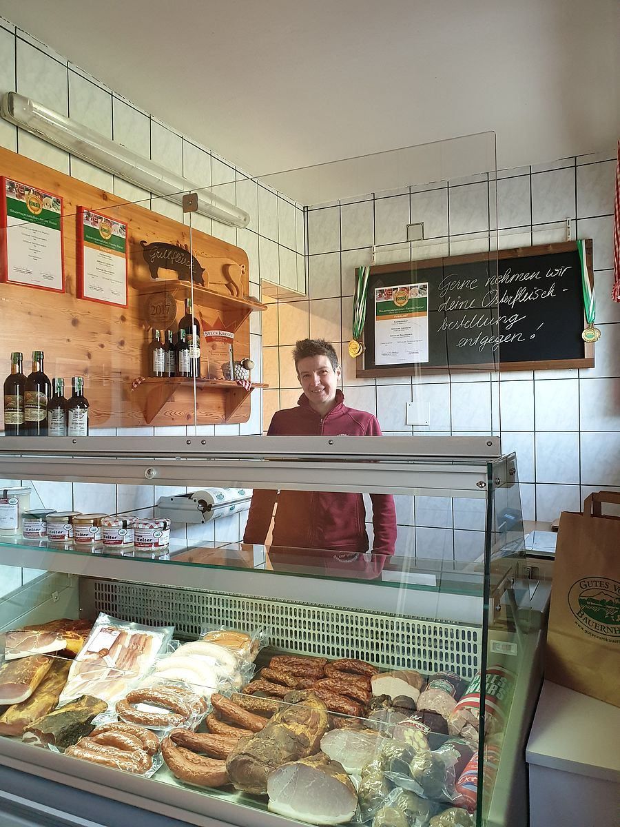 Ein Mann steht hinter einer Glasvitrine, die mit verschiedenen Fleischsorten und Würsten gefüllt ist, und lächelt. Über ihm befindet sich eine Tafel mit einem deutschen Spruch und zwei Medaillen. Holzregale halten Flaschen und Auszeichnungen, während ein brauner Papierbeutel rechts neben ihm steht.