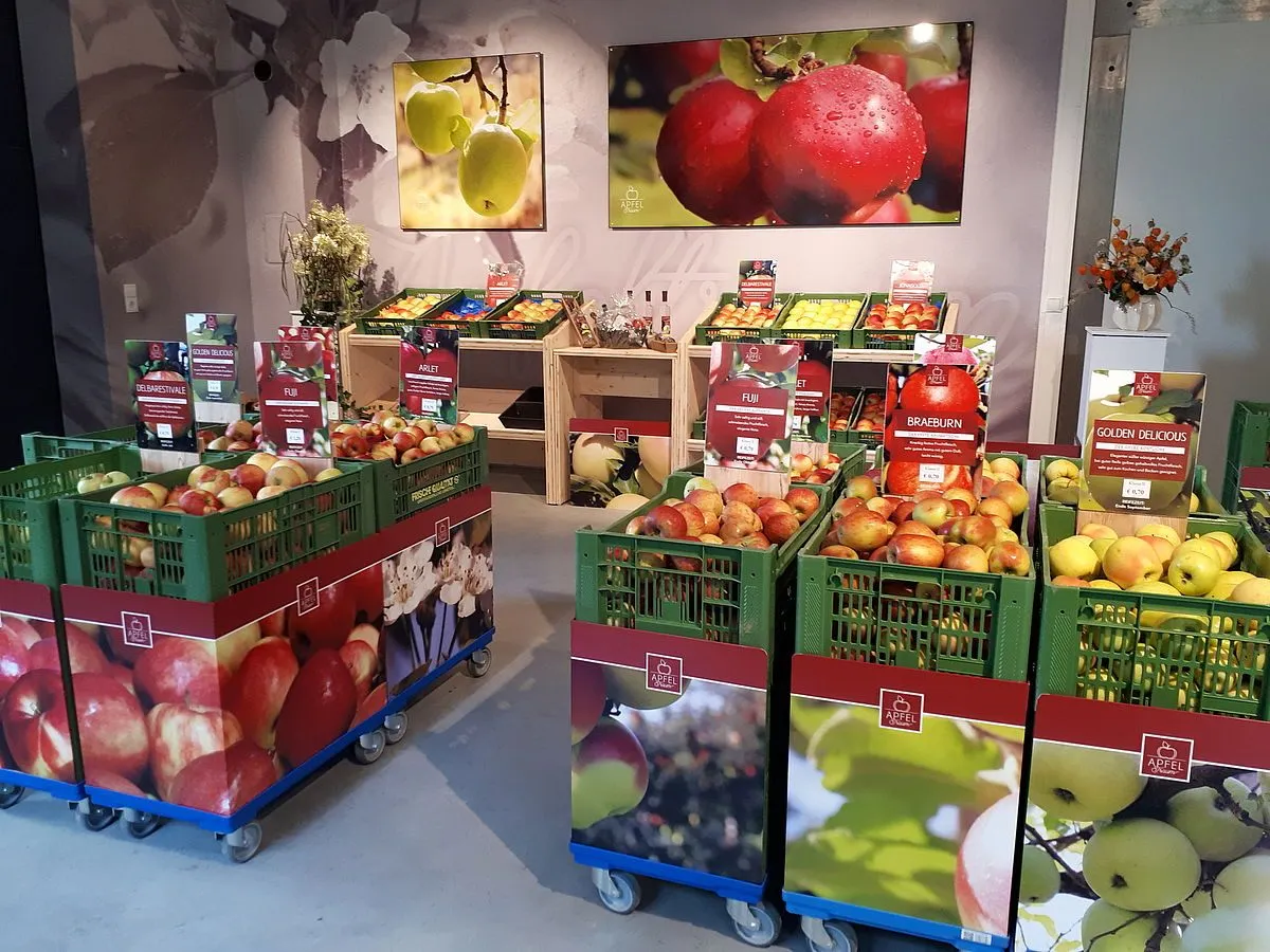Ein Laden gefüllt mit verschiedenen Kisten voller Äpfel, die prominent Schilder für Fuji und Braeburn anzeigen. An der Wand hängen große Gemälde von Äpfeln, eines zeigt einen roten Apfel mit Regentropfen.