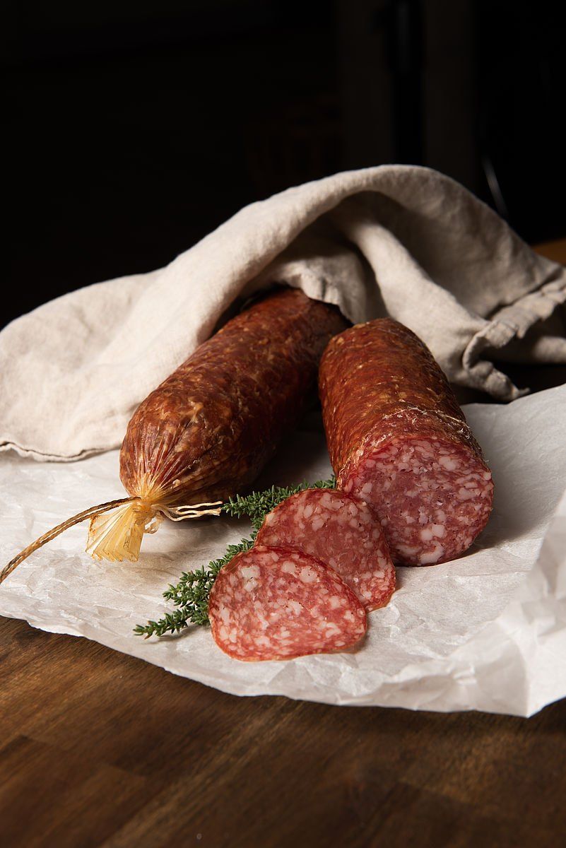 Zwei Scheiben Salami liegen auf einem Stück Papier, eine teilweise von einem Tuch bedeckt. Daneben befindet sich ein Bündel Salami mit einem gelben Faden. Ein Rosmarinzweig ist ebenfalls sichtbar.