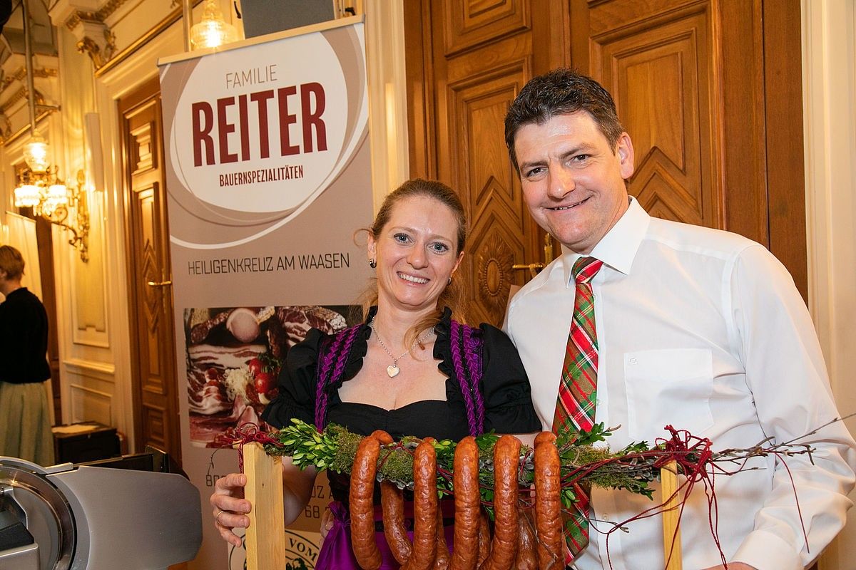 Ein Mann und eine Frau stehen lächelnd neben einem Banner für Familie Reiter und halten eine Girlande aus Würsten. Der Mann trägt ein weißes Hemd und eine Krawatte, während die Frau ein traditionelles Kleid trägt. Sie stehen vor Holztüren.