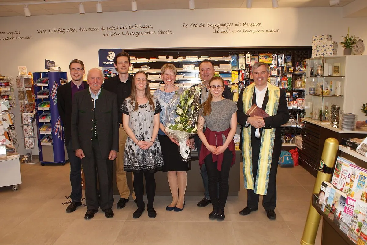 Eine Gruppe von Menschen steht in einem Raum mit einem deutschen Text an der Wand. Die Frau in der Mitte hält einen Blumenstrauß. Der Mann rechts trägt einen Umhang und hält ein Buch. Hinter ihnen sind Regale mit Produkten.
