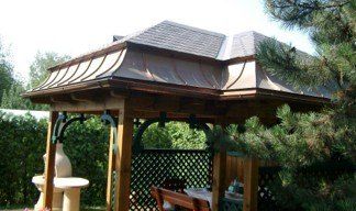 Ein hölzerner Pavillon mit Schieferdach und Gitterzaun steht in einem Garten, umgeben von Bäumen und Grün. Im Inneren befinden sich ein Tisch und Stühle.