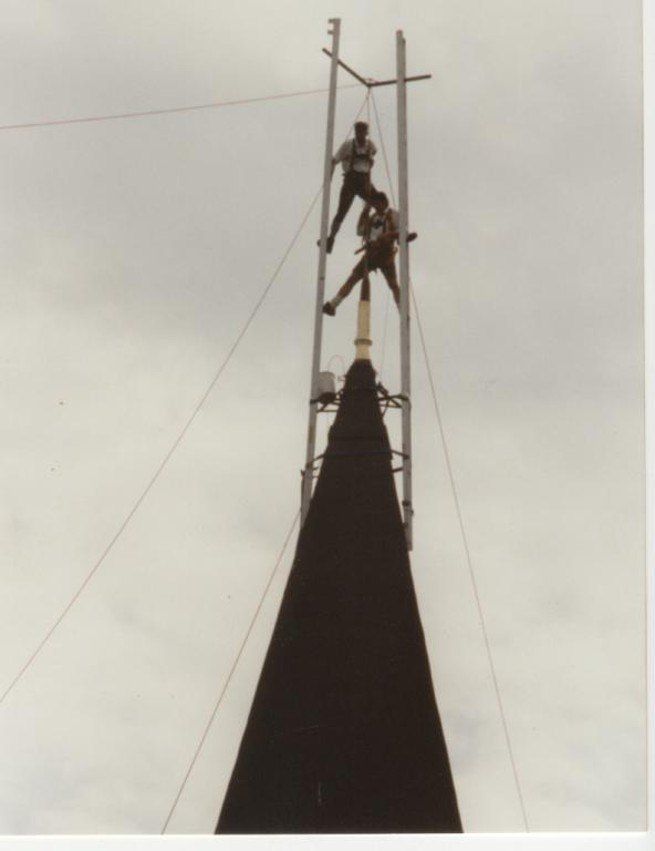 Zwei Personen klettern einen hohen Turm hoch und tragen Gurte. Der Turm ist aus Metall und hat daran befestigte Drähte.