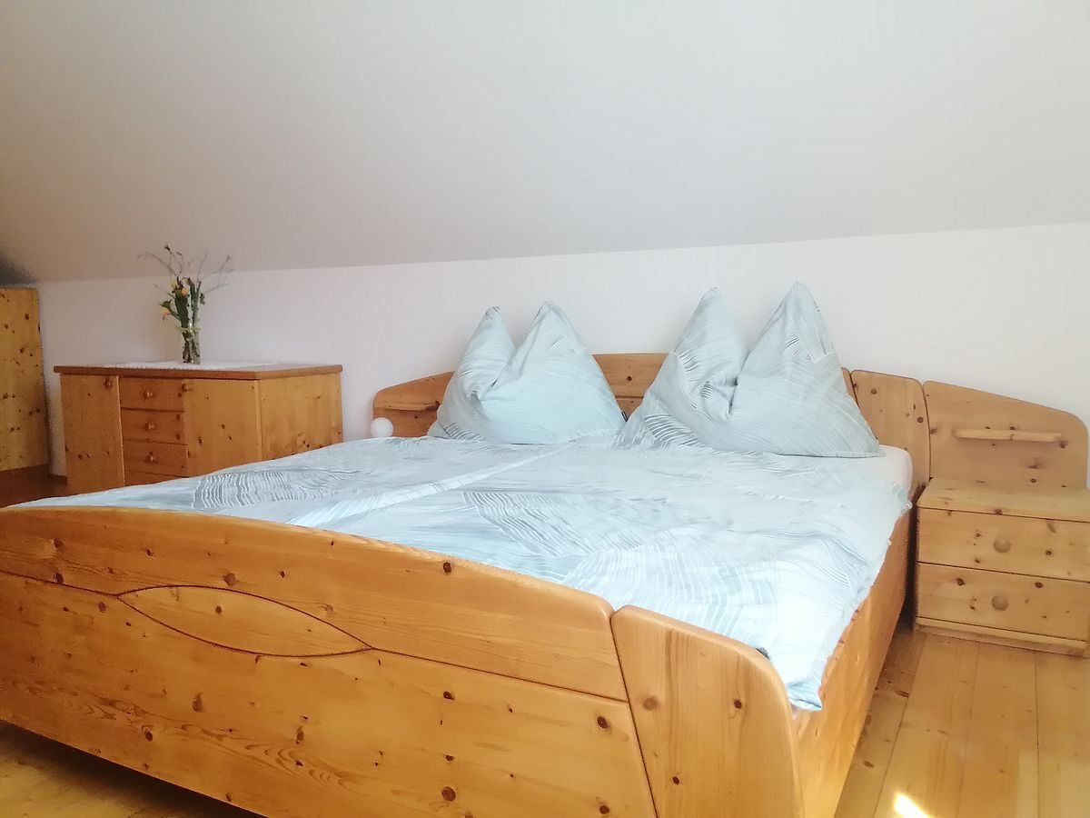 Ein gemütliches Schlafzimmer mit einem Holzbett und einer Kommode, geschmückt mit einer Blumenvase. Das Bett hat eine hellblaue Decke und Kissen, und der Raum hat eine weiße Wand und einen Holzboden.