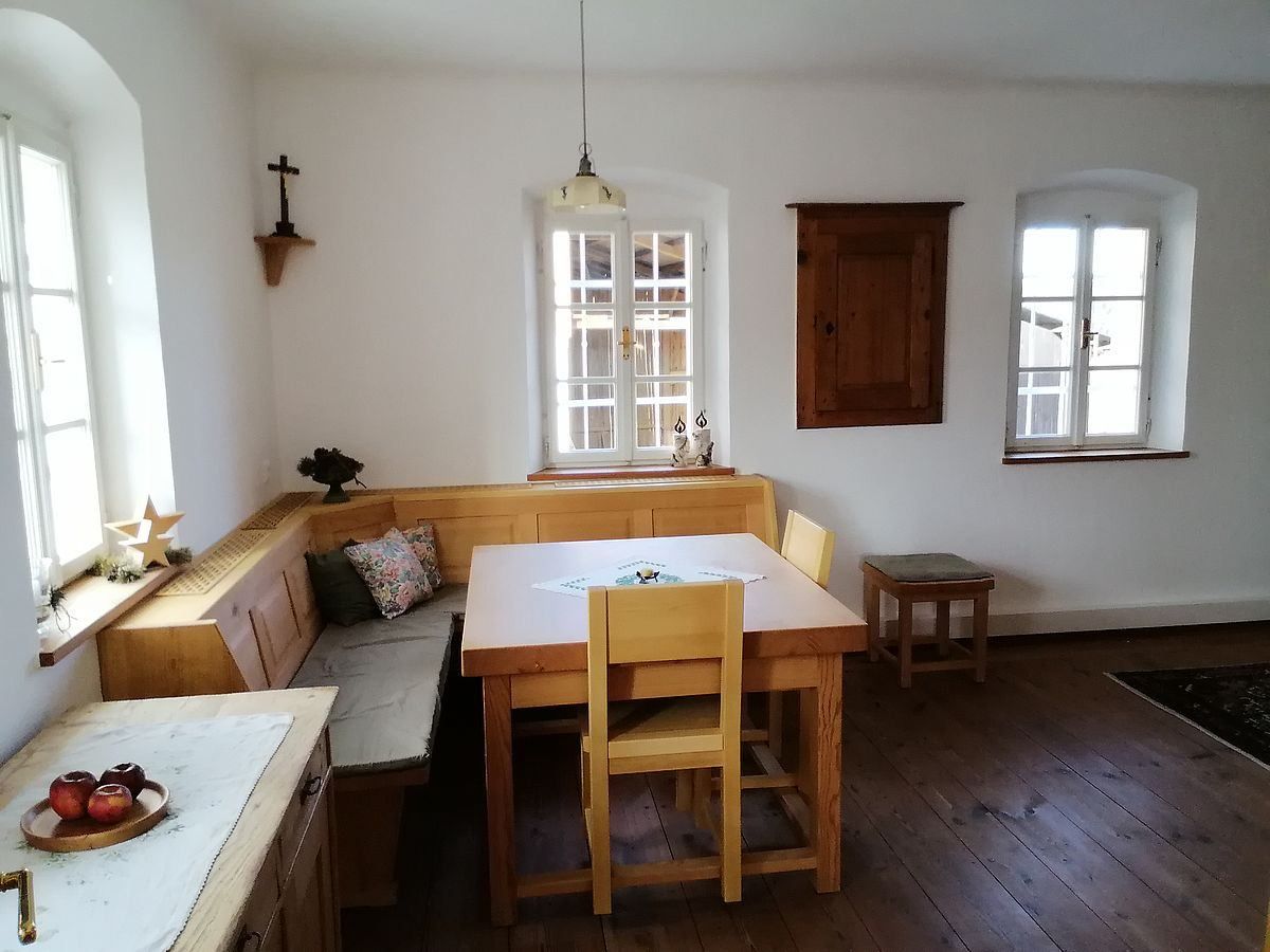 Ein Essbereich mit einem Holztisch und Stühlen, einer Eckbank mit Kissen, einer weißen Tischdecke und zwei Fenstern. Ein Holzschrank ist an der Wand montiert.