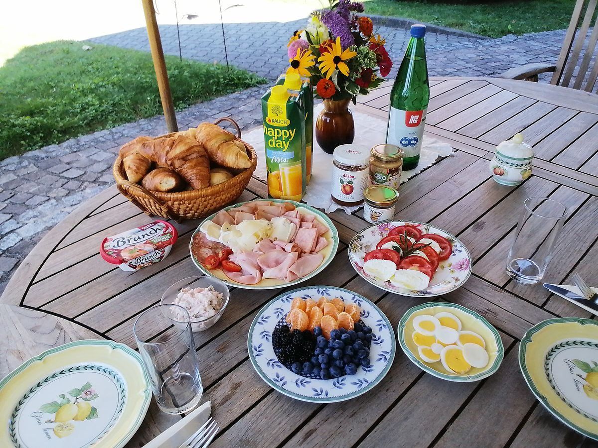 Ein Frühstückstisch im Freien mit Croissants, Eiern, Obst und Getränken. Eine Vase mit Blumen und eine Flasche Saft stehen auf dem Tisch.