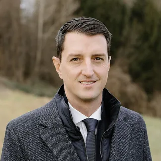 Ein Mann mit kurzen Haaren, lächelnd, trägt einen grauen Anzug und eine schwarze Krawatte. Er steht auf einem Rasenfeld mit Bäumen im Hintergrund.