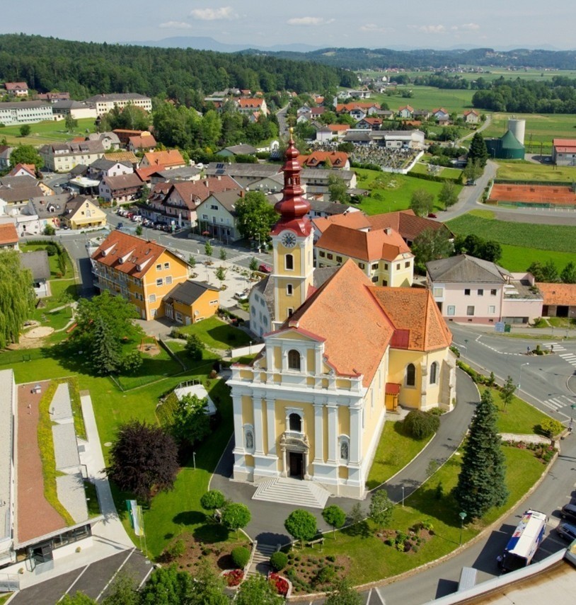 Ein Luftbild einer Stadt mit zahlreichen Gebäuden, einer prominenten Kirche und einer umliegenden Landschaft aus Bäumen, Feldern und Straßen.