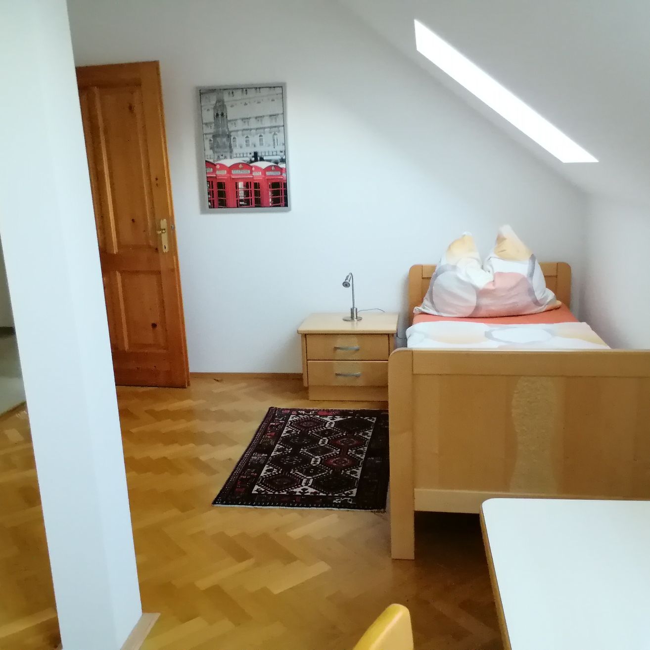 Ein gemütliches Schlafzimmer mit Holzboden, einem Bett, einem Nachttisch mit Lampe und einem Bilderrahmen an der Wand. Der Raum hat ein Dachfenster und eine Holztür.