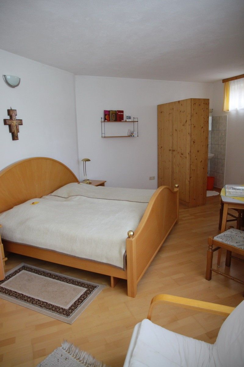 Ein Schlafzimmer mit einem Doppelbett und einem Nachttisch, einem Holzschrank, einer Lampe, einem Teppich und einem Regal mit Büchern. An der Wand befinden sich ein Kreuz und eine Lampe.