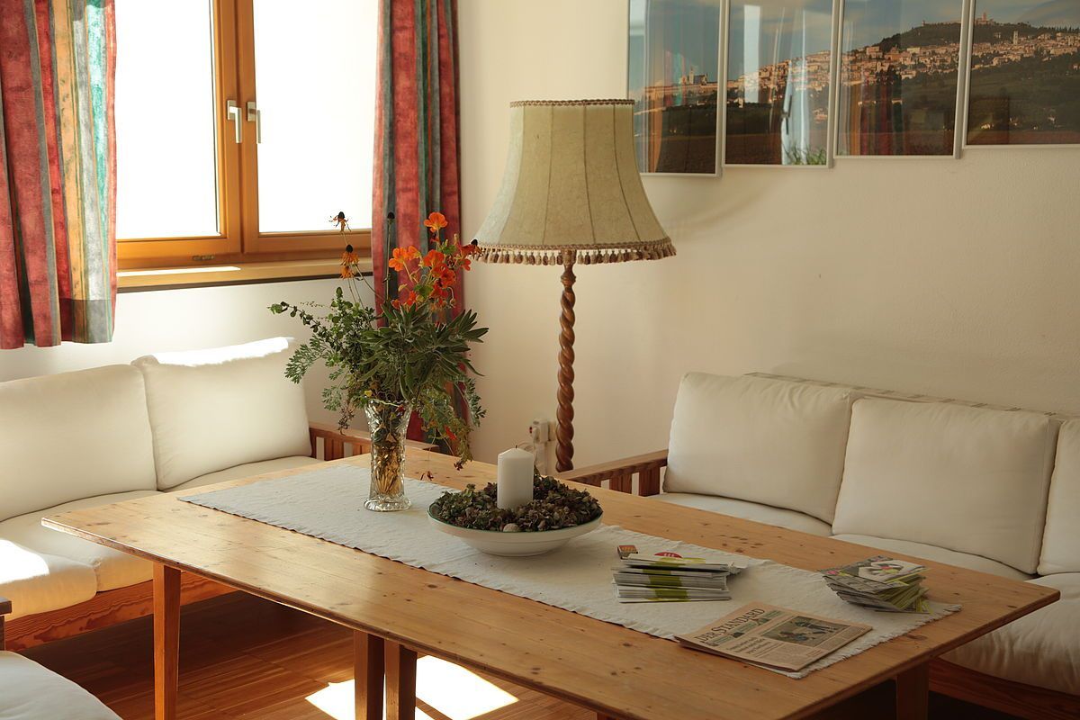 Ein gemütliches Zimmer mit einem Holztisch, einer Lampe und einer Couch mit weißen Kissen. Auf dem Tisch stehen ein Blumenvase, eine Kerze und einige Zeitschriften.