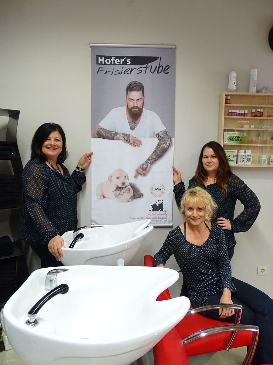 Drei Frauen in einem Salon halten ein Poster eines bärtigen Mannes mit Tattoos. Dahinter befindet sich ein Regal mit verschiedenen Produkten. Im Vordergrund stehen zwei Waschbecken.