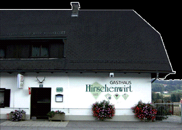 Ein weißes Gebäude mit einem schwarzen Dach. Es zeigt ein Schild mit der Aufschrift Gasthaus Hirschenwirt über einer Tür. Ein Hirschkopf ist über dem Eingang montiert. Es gibt Blumen in Pflanzgefäßen auf beiden Seiten der Tür.