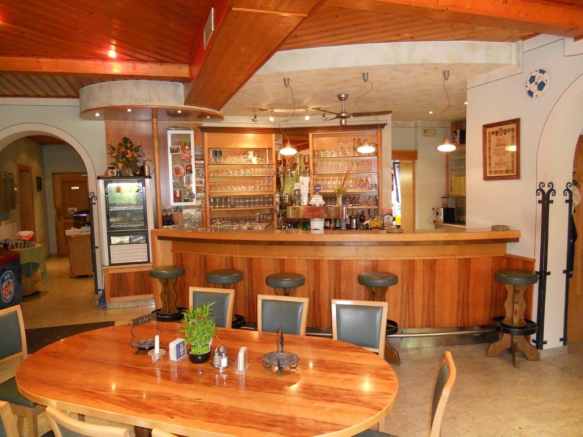 Eine gemütliche Bar mit Holztheken, Hockern und Tischen. Regale gefüllt mit Gläsern und Flaschen. Eine Holzdecke und hängende Lampen.