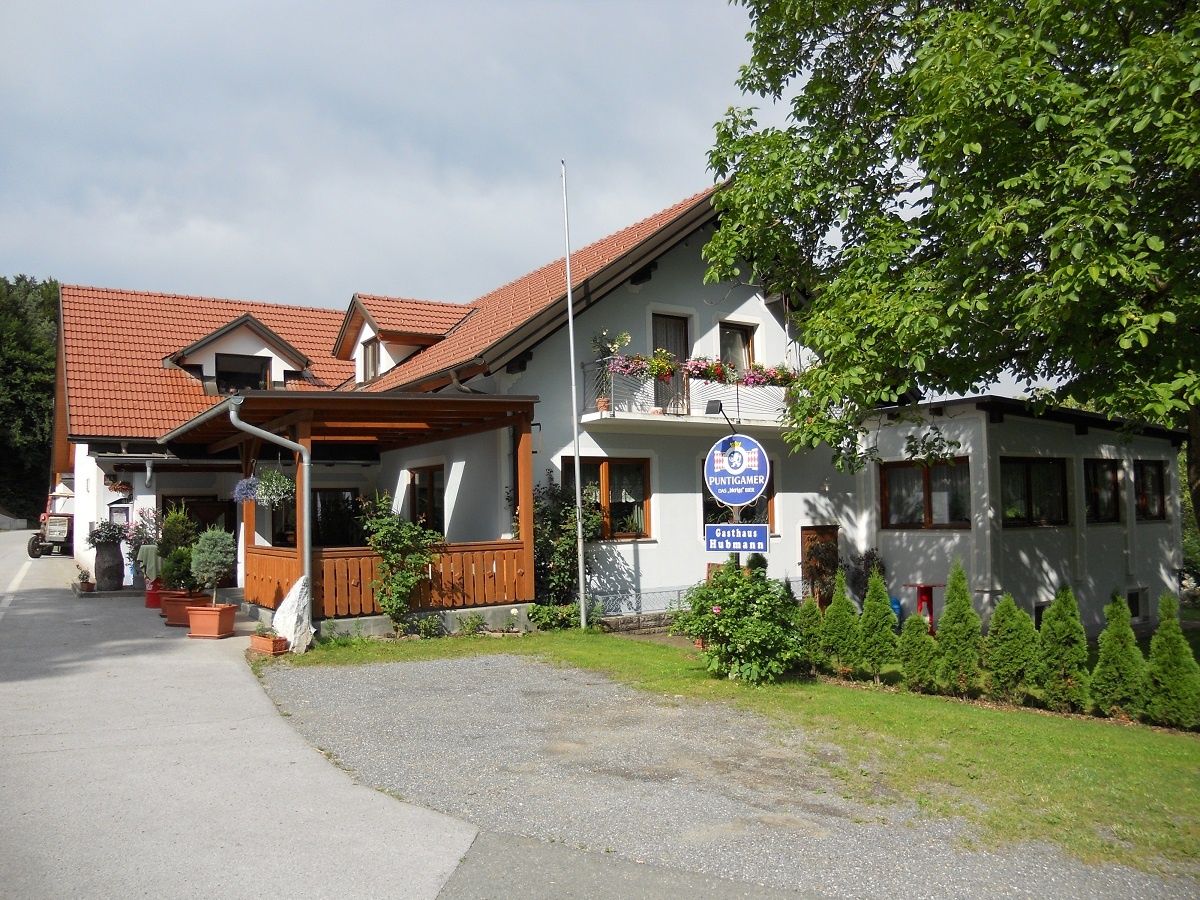 Ein weißes Haus mit rotem Dach, Balkonen und einem Schild für Puntigamer. Die Vorderseite hat eine Veranda mit Topfpflanzen. Bäume und Sträucher umgeben das Haus.