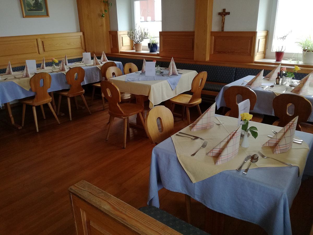 Ein Restaurantinnenraum mit mehreren Tischen, die zum Essen gedeckt sind. Jeder Tisch hat eine blaue Tischdecke, ein weißes Serviettentuch und eine gefaltete Menükarte. Besteck wie Gabeln, Messer und Löffel sind ordentlich angeordnet. Es gibt Holzstühle und Bänke. Der Raum hat Holzwände und -böden und ein Fenster mit einem Kreuz auf der Fensterbank.