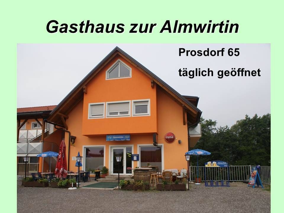 Das Gasthaus zur Almwirtin ist ein leuchtend orangefarbenes Gebäude mit Prossdorf 65 oben drauf. Es hat mehrere Glastüren und Fenster. Draußen stehen Tische und Stühle mit Sonnenschirmen.