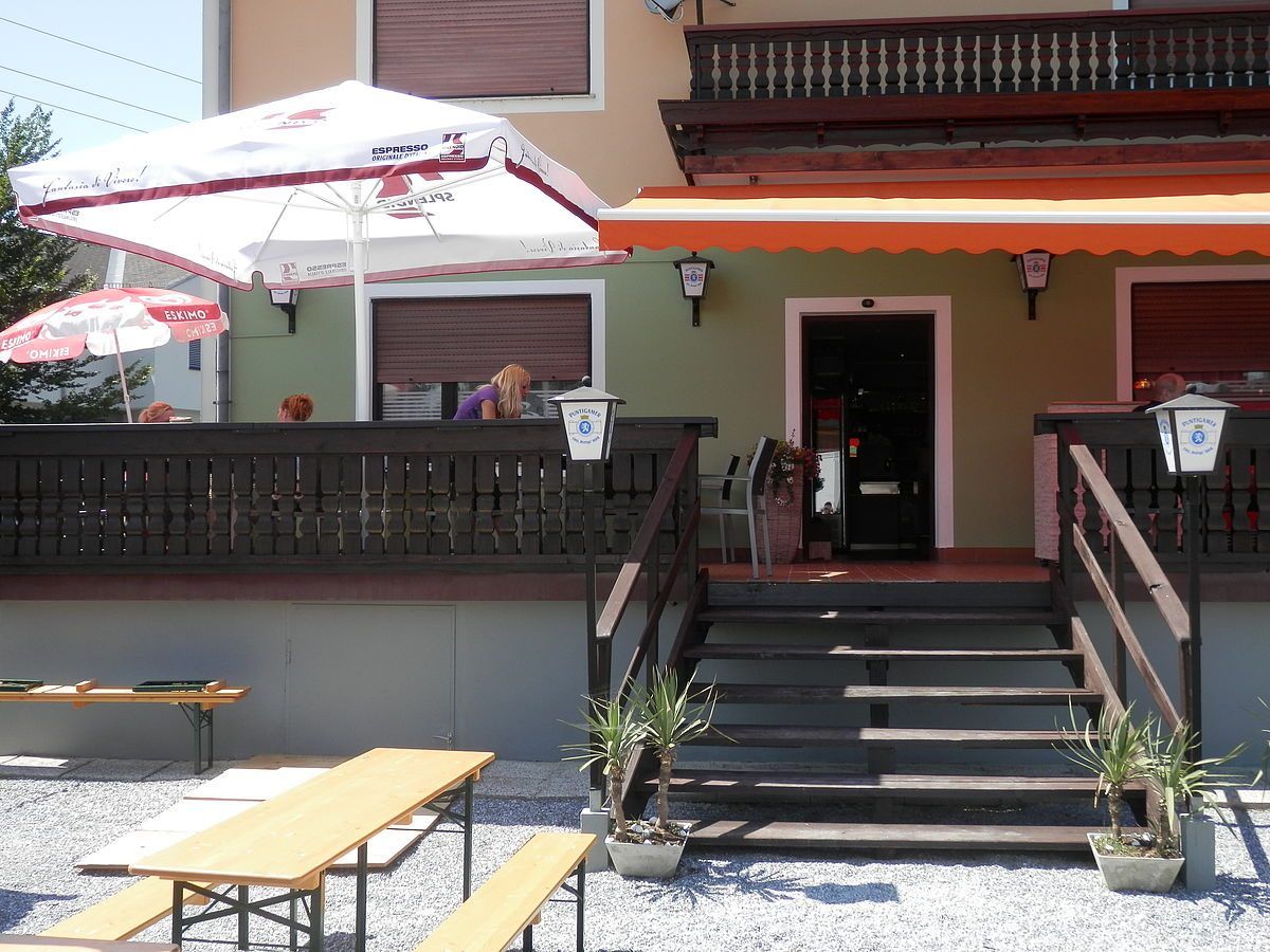 Das Äußere eines Cafés mit einem Holztisch, einem Stuhl und einem Schirm. Ein Vordach bedeckt den Eingang. Zwei Frauen sitzen auf dem Balkon.