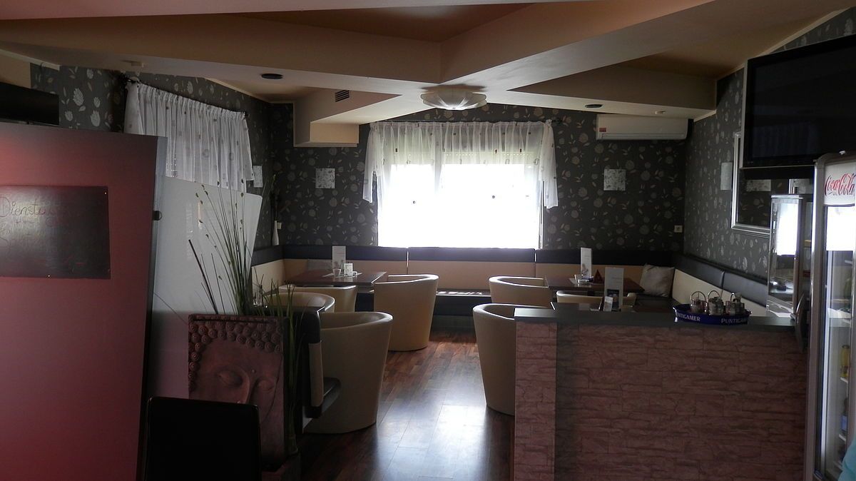 Ein Raum mit einer beige und dunkel gemusterten Wand, einem Fenster mit weißen Vorhängen, einer Decke mit einer Lampe und einem Holzboden. Es gibt runde Stühle um einen Tisch mit Tassen und eine Theke mit einem Eimer.