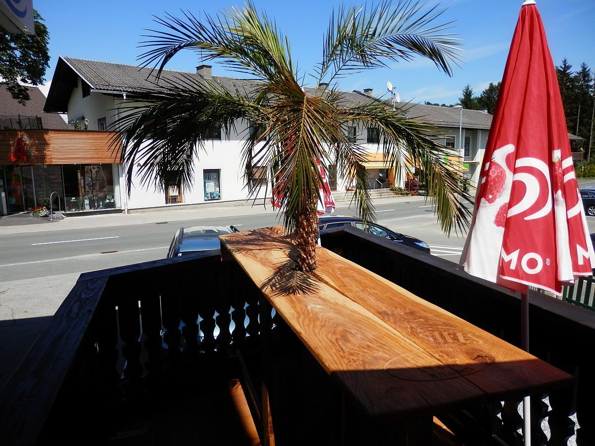 Ein Balkonblick mit einer Palme auf einem Holztisch. Zwei Autos sind auf der Straße geparkt, und ein Gebäude ist im Hintergrund. Der Himmel ist klar.