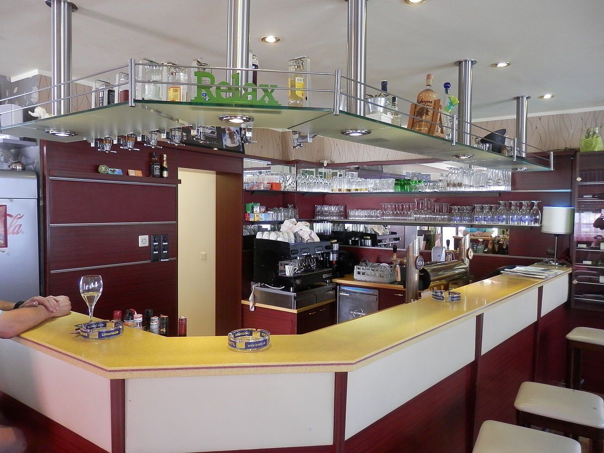 Eine Bar mit gelben Arbeitsplatten und Regalen, die mit verschiedenen Likörflaschen bestückt sind. Die Bar hat ein hängendes Regal mit dem Wort 'Relax'. Verschiedene Gläser und Gegenstände sind in den Regalen.