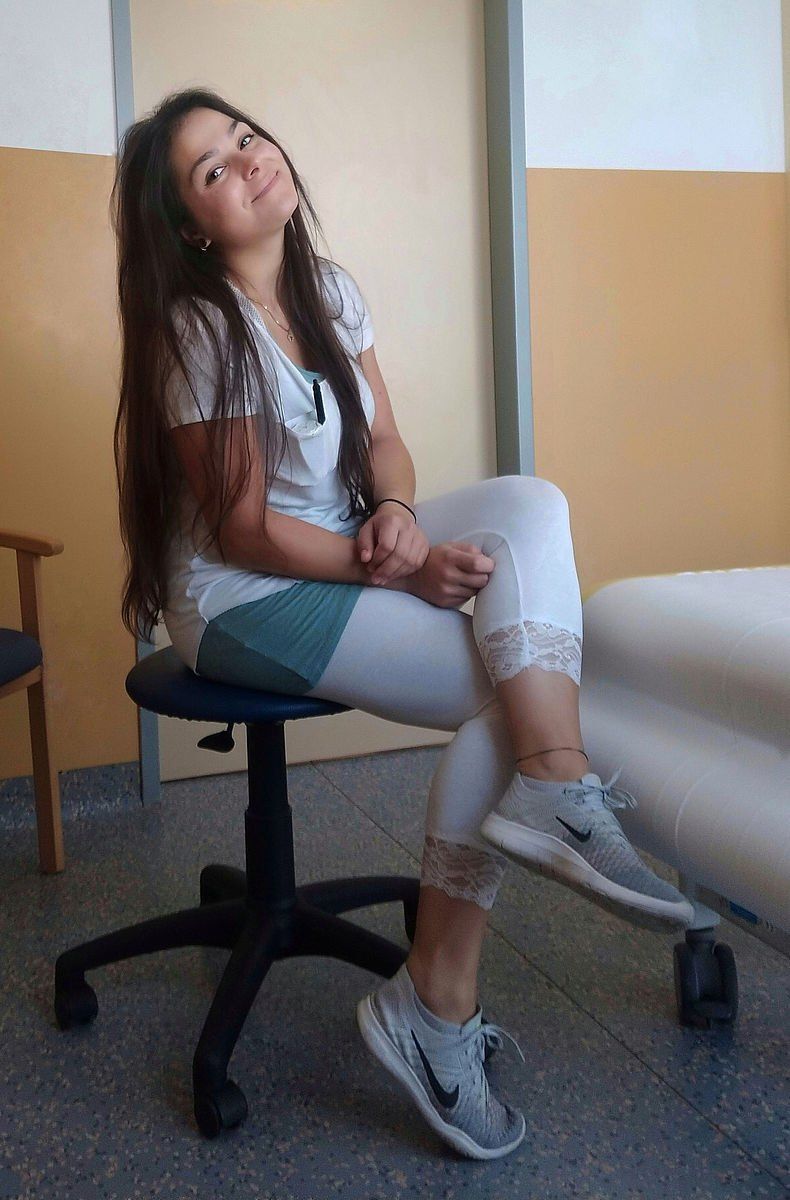 Eine junge Frau sitzt auf einem Stuhl in einem Raum. Sie trägt ein weißes Shirt, bunte Leggings und Nike-Sneaker. Sie lächelt und wirkt entspannt.