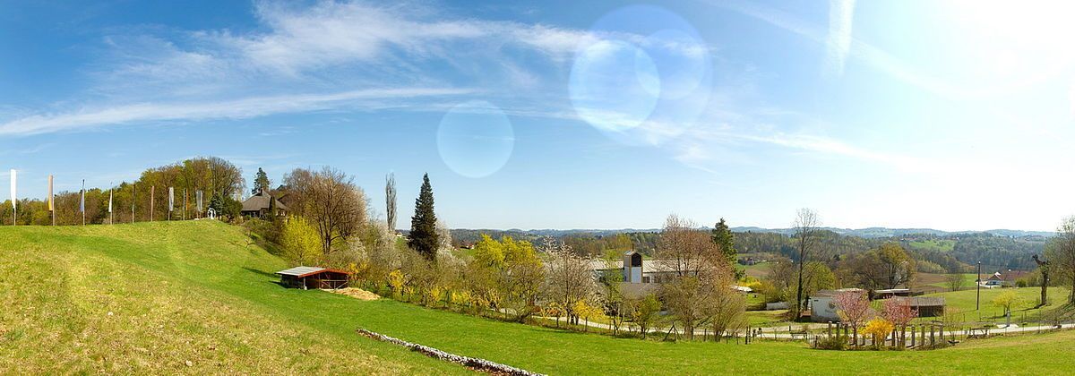 Eine malerische ländliche Landschaft mit einem grünen Hügel, Häusern und Bäumen unter einem klaren blauen Himmel. Im Vordergrund befindet sich ein kleiner Schuppen und ein gewundener Weg.