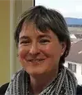 Eine Frau mit kurzen Haaren und einem grauen Schal lächelt in die Kamera vor einem Fenster mit Blick auf Gebäude.
