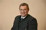 Ein Mann in einem schwarzen Anzug und einer Krawatte lächelt in die Kamera und steht vor einer beigefarbenen Wand.