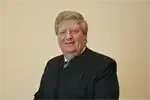 Ein Mann in einem schwarzen Anzug, der eine Brille trägt, lächelt und posiert für ein Foto vor einer hellen Wand.