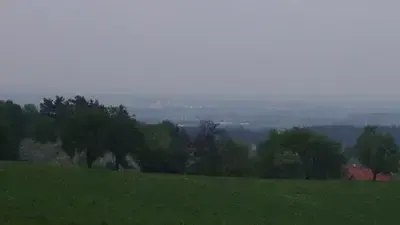 Ein Blick von einem Hügel zeigt ein weites grünes Feld mit Bäumen und einem roten Gebäude in der Ferne. Der Himmel ist bewölkt.