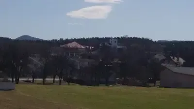 Eine ländliche Landschaft mit einem grasigen Feld, einigen Häusern und Bäumen. In der Ferne gibt es Berge und einen klaren blauen Himmel.