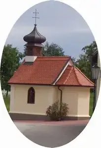 Eine kleine Kapelle mit rotem Dach und einem Kreuz oben drauf. Sie ist von Grün umgeben und hat ein Fenster mit einer Statue darin.