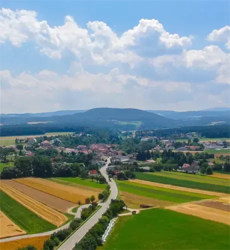 Luftaufnahme einer ländlichen Landschaft mit Feldern, einer kurvenreichen Straße und einem kleinen Dorf, umgeben von Bäumen und Bergen unter einem bewölkten Himmel.
