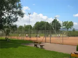 Ein Tennisplatz mit einigen Personen, die Tennis spielen, umgeben von einem Metallzaun, mit Bänken und Bäumen in der Umgebung.