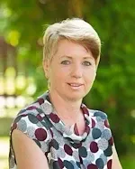 Eine Frau mit kurzen, blonden Haaren lächelt vor einem unscharfen grünen Hintergrund und trägt ein gepunktetes Oberteil.