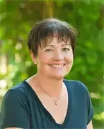 Eine Frau mit kurzen Haaren lächelt und trägt ein blaues Shirt. Sie hat eine Halskette und steht vor grünen Bäumen.
