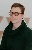 Eine Person mit kurzen Haaren, die eine Brille und ein schwarzes Rollkragenshirt trägt, steht vor einer Wand mit dreieckigen Mustern.