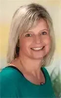 Eine Frau mit kurzen blonden Haaren, die ein blaues Hemd trägt, lächelt und in die Kamera schaut. Sie trägt Ohrringe und eine Halskette.