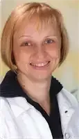 Eine Frau mit kurzen blonden Haaren, die eine weiße Laborkittel mit schwarzem Kragen trägt, lächelt fröhlich in die Kamera. Sie steht in einem Raum mit einer hellen Wand hinter ihr.