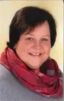 Eine lächelnde Frau mit kurzen schwarzen Haaren und einem roten Schal posiert für ein Foto vor einer hellen Wand.