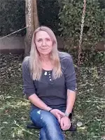 Eine Frau mit blonden Haaren sitzt auf einer Schaukel im Garten, trägt ein graues Shirt und Jeans und lächelt.