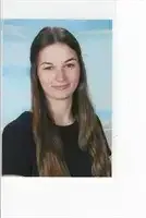 Eine lächelnde Frau mit langen, gewellten Haaren posiert für ein Foto vor einem unscharfen Hintergrund. Sie trägt ein schwarzes Oberteil und schaut geradeaus.